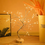 Decorative LED tree on a table with a warm glow, next to a vase with flowers and a framed picture.